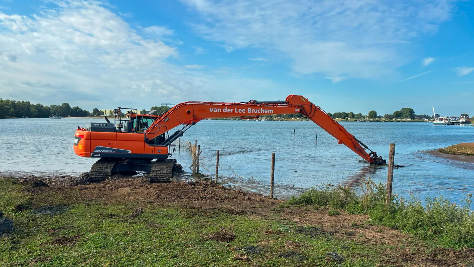 Overzichtsfoto van baggerwerkzaamheden aan een brede watergang. Een rupskraan van van der Lee Bruchem werkt vanuit het water aan het verwijderen van slib. De omgeving bestaat uit groen, een zandstrandje en een industriële haven.