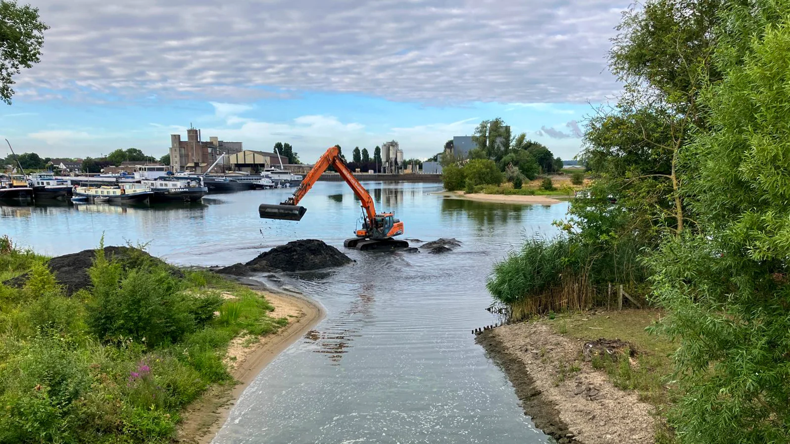 Overzichtsfoto van baggerwerkzaamheden aan een brede watergang. Een rupskraan van van der Lee Bruchem werkt vanuit het water aan het verwijderen van slib. De omgeving bestaat uit groen, een zandstrandje en een industriële haven.