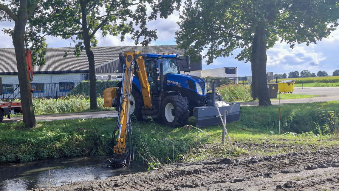 New Holland-tractor met maaikorf van Van der Lee Bruchem aan het werk langs een watergang.