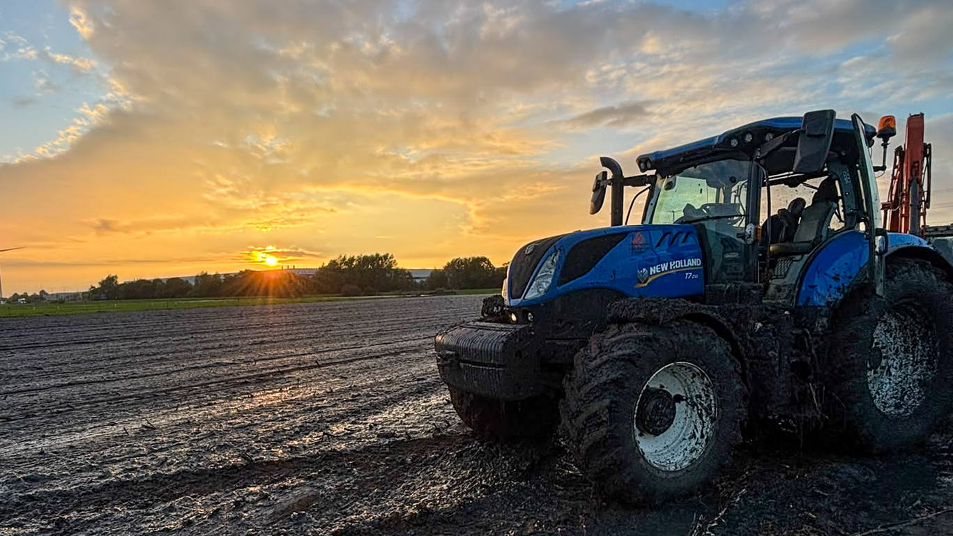 Blauwe New Holland-tractor van Van der Lee Bruchem op een drassige akker bij zonsondergang.