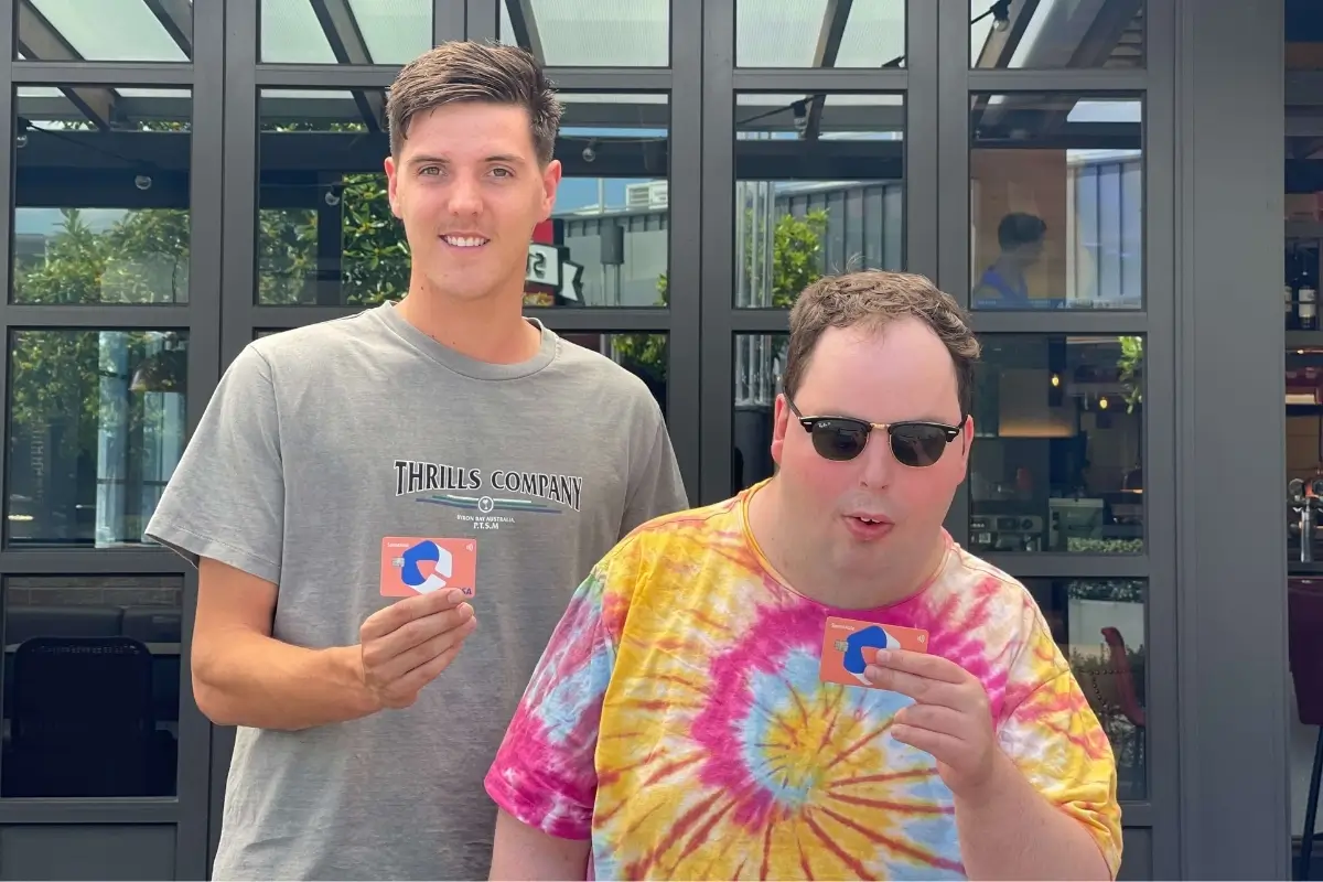 Two men, one wearing a grey t-shirt and the other in a colorful tie-dye shirt and sunglasses, smile and proudly display their SpendAble Visa cards outside a modern cafe. The image highlights inclusive finance and how a money management app can empower users of all abilities to take control of their spending.