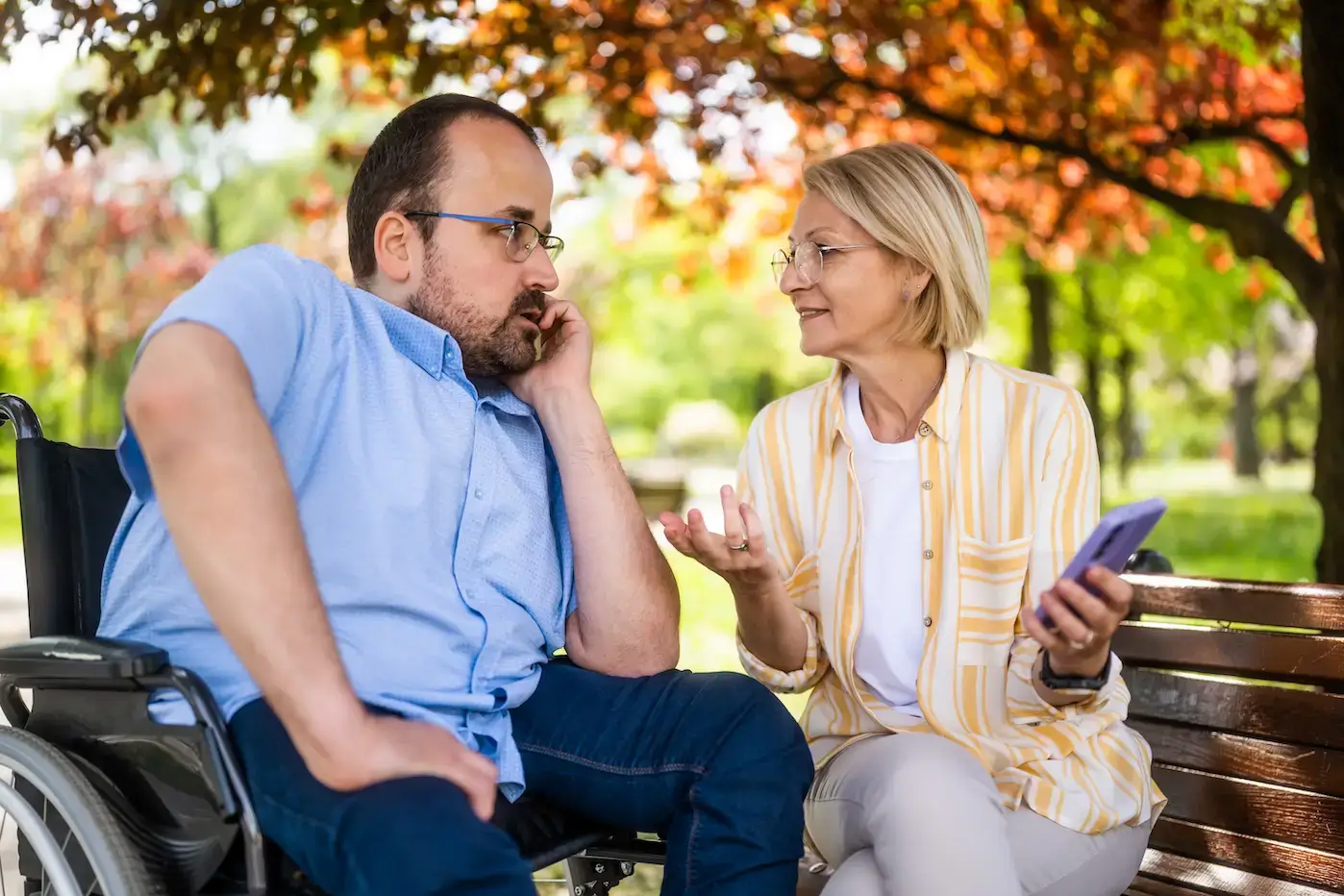A woman holding a smartphone sits on a park bench talking with a man in a wheelchair, showing support and guidance with using a money management app in Australia.