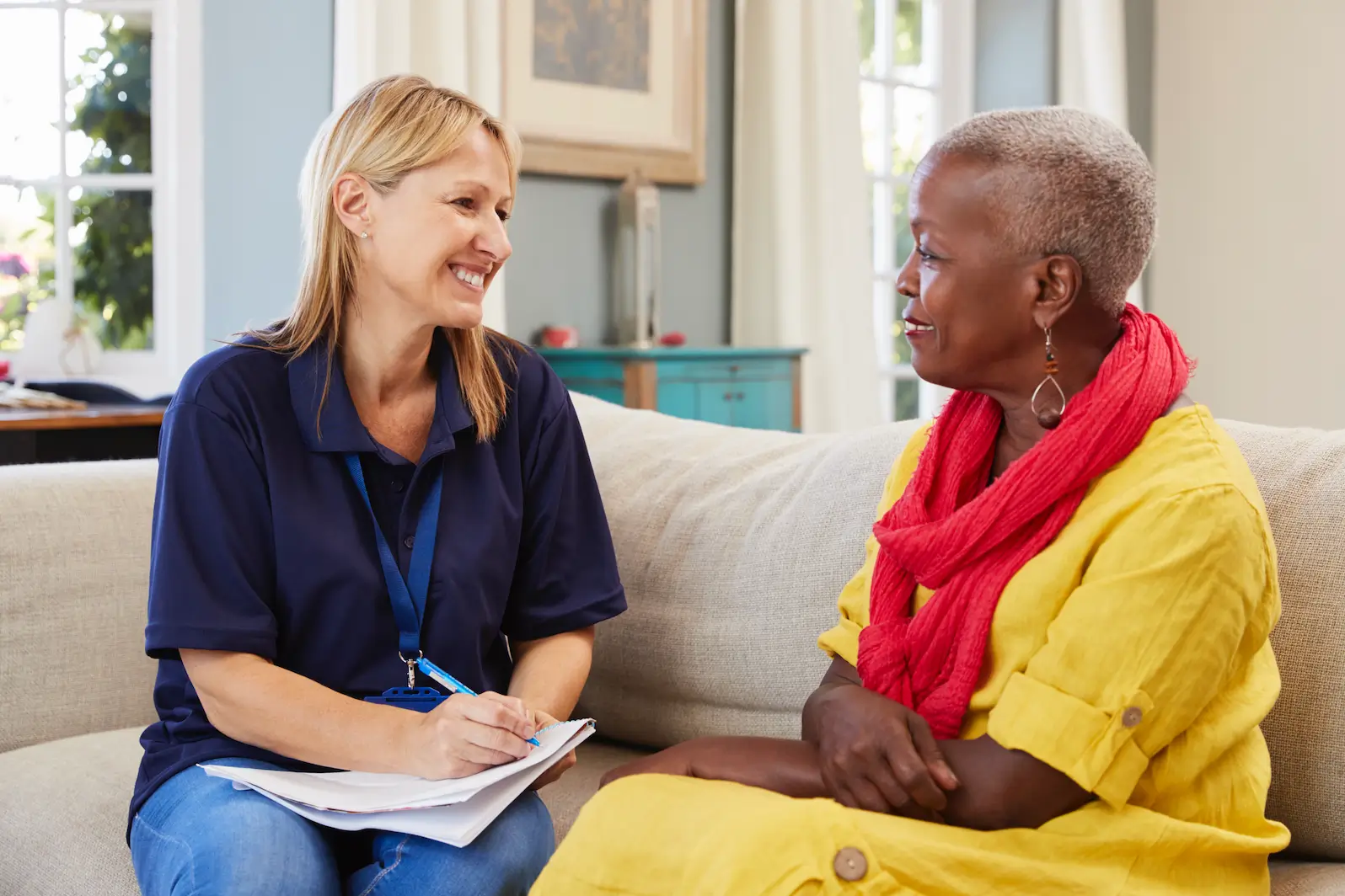 Two women smiling and talking on a couch, with one writing notes on a clipboard, showing how an app for support workers helps build trust and communication.