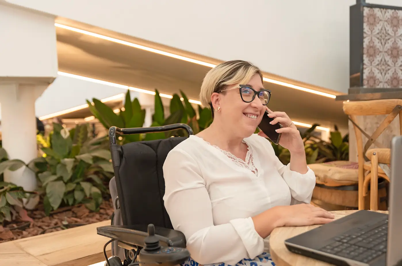 A smiling woman in a wheelchair talks on her phone while working on her laptop at a café, showing independence and confidence related to understanding what does allocated funds mean NDIS.