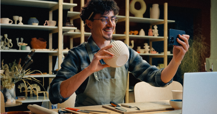 A man in an apron holds up a ceramic cup as he takes a selfie with his phone.