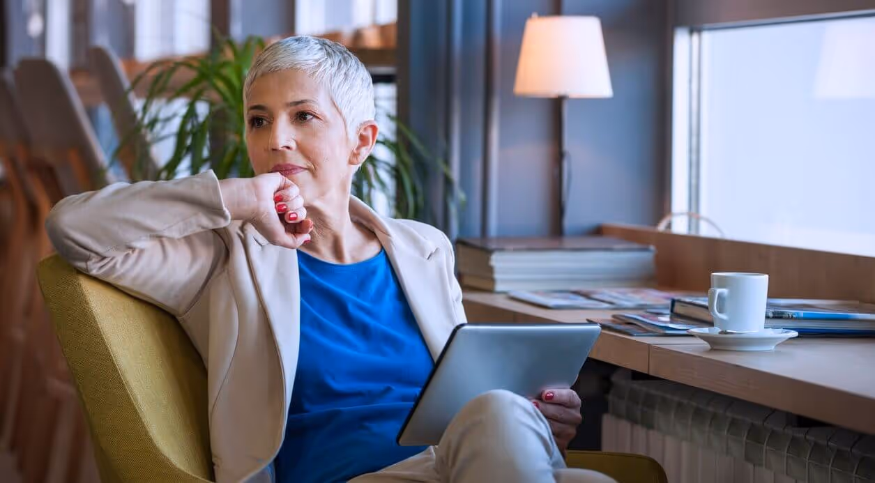 A thoughtful senior woman, an aspiring business owner, holds a tablet while considering the process of starting a new business.