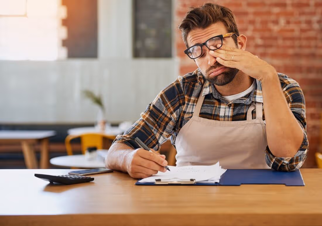 A tired, stressed small business owner in an apron rubs his eye while working on financial papers and a calculator.