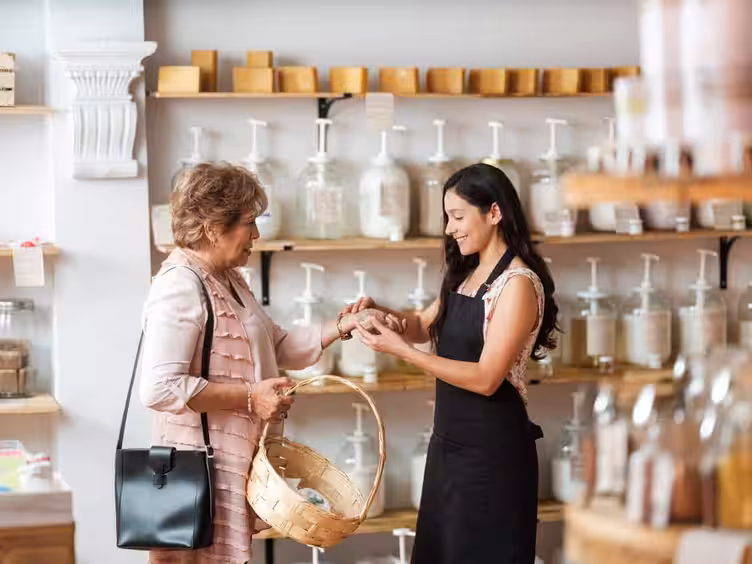 A smiling small business owner in an apron shows a product to an older customer holding a basket in a modern, eco-friendly store.