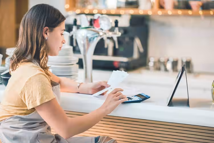 A small business owner in an apron calculates gross revenue using receipts and a calculator, managing finances for her business.