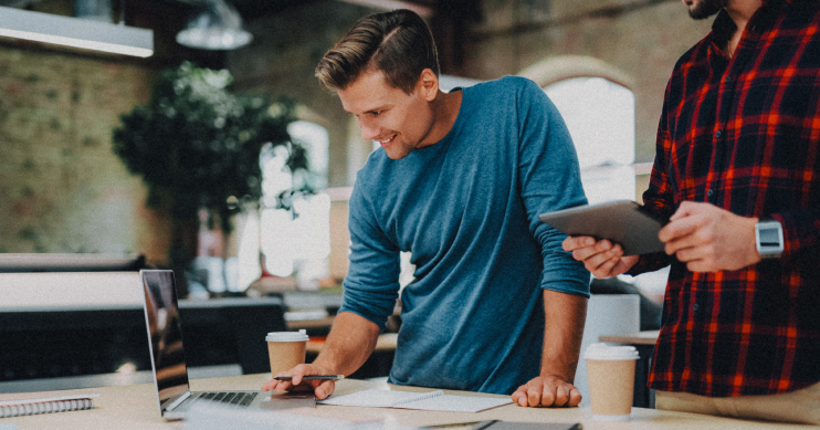 A man smiles as he types on a laptop, surrounded by papers and coffee cups.