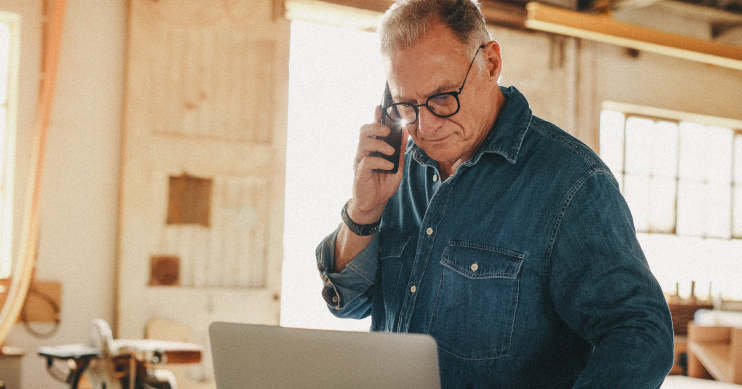 a man in a blue denim shirt looks down at a laptop while holding a phone to his ear.