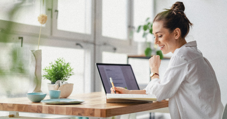 A woman with a laptop open in front of her writes in a notebook.