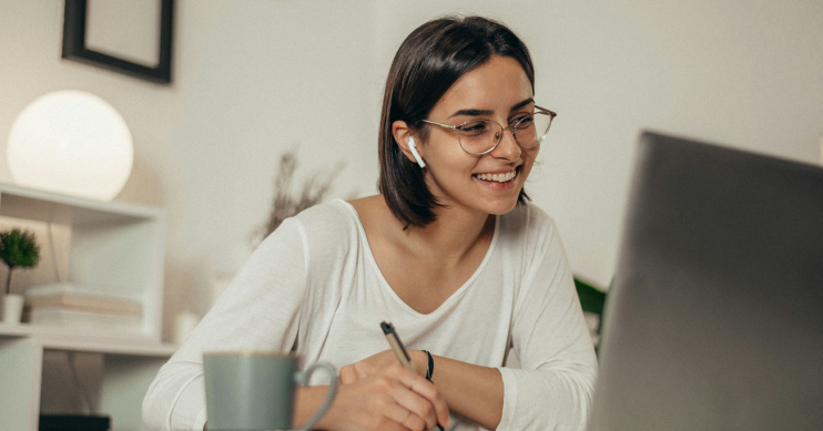 A woman smiles at her laptop as she writes something on a notepad.