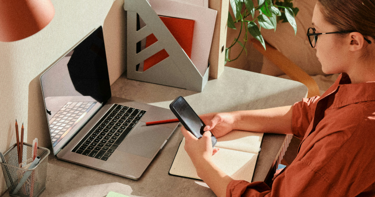 A woman looks at her phone while seated at her desk, in front of a laptop.