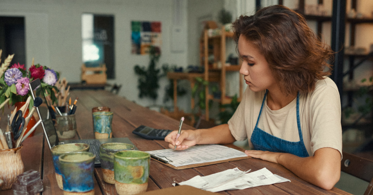 A woman in a work apron sits at a worktable writing with a pen and paper.