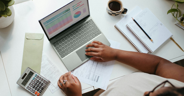 A person works at a laptop, surrounded by papers, and a calculator.
