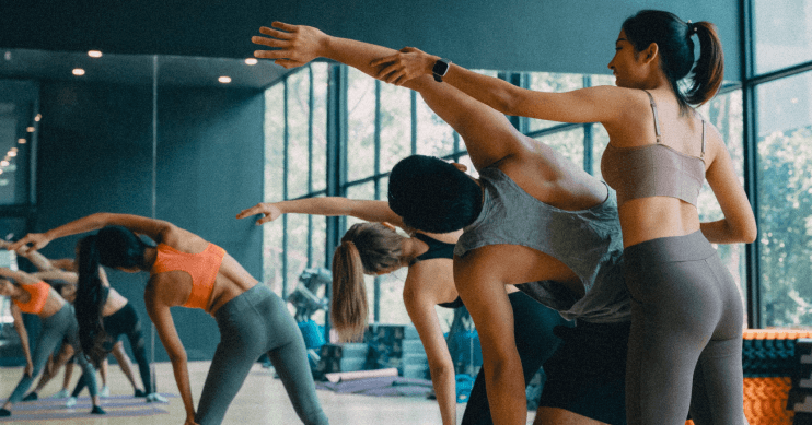 Women stretch with the help of an instructor in a gym studio.