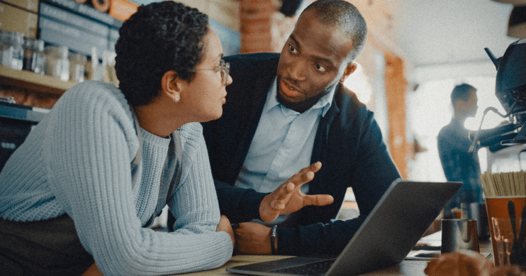 A woman leans on a table in front of a laptop, listening to a man in conversation.