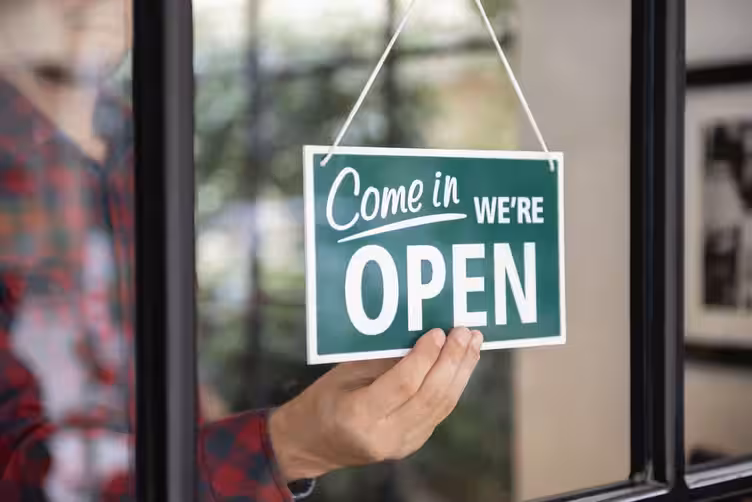 A hand turns a green 'Come in WE'RE OPEN' sign in a glass door, symbolizing a turnkey business ready for immediate operation.