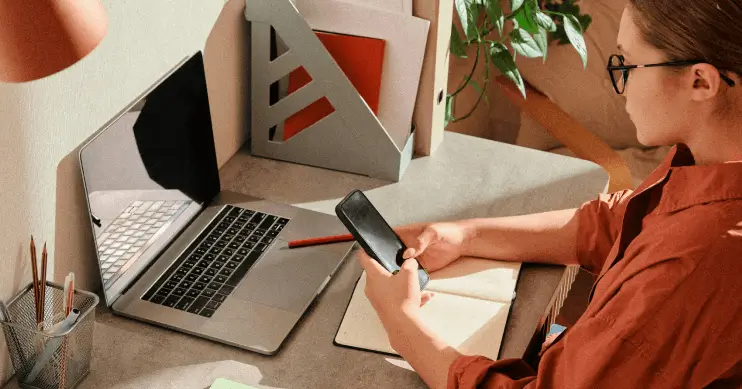 A woman looks at her phone while seated at her desk, in front of a laptop.