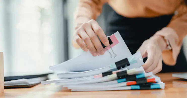 A woman lifts a bound stack of paper off a pile of other papers.