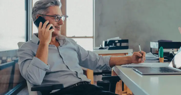 A man sits at a desk looking at a laptop while on an office phone.
