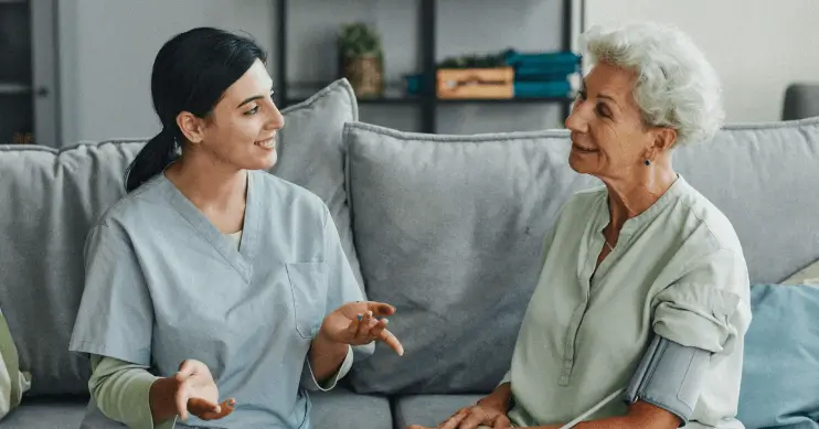 A woman in nurses scrubs sits talking with an elderly patient.
