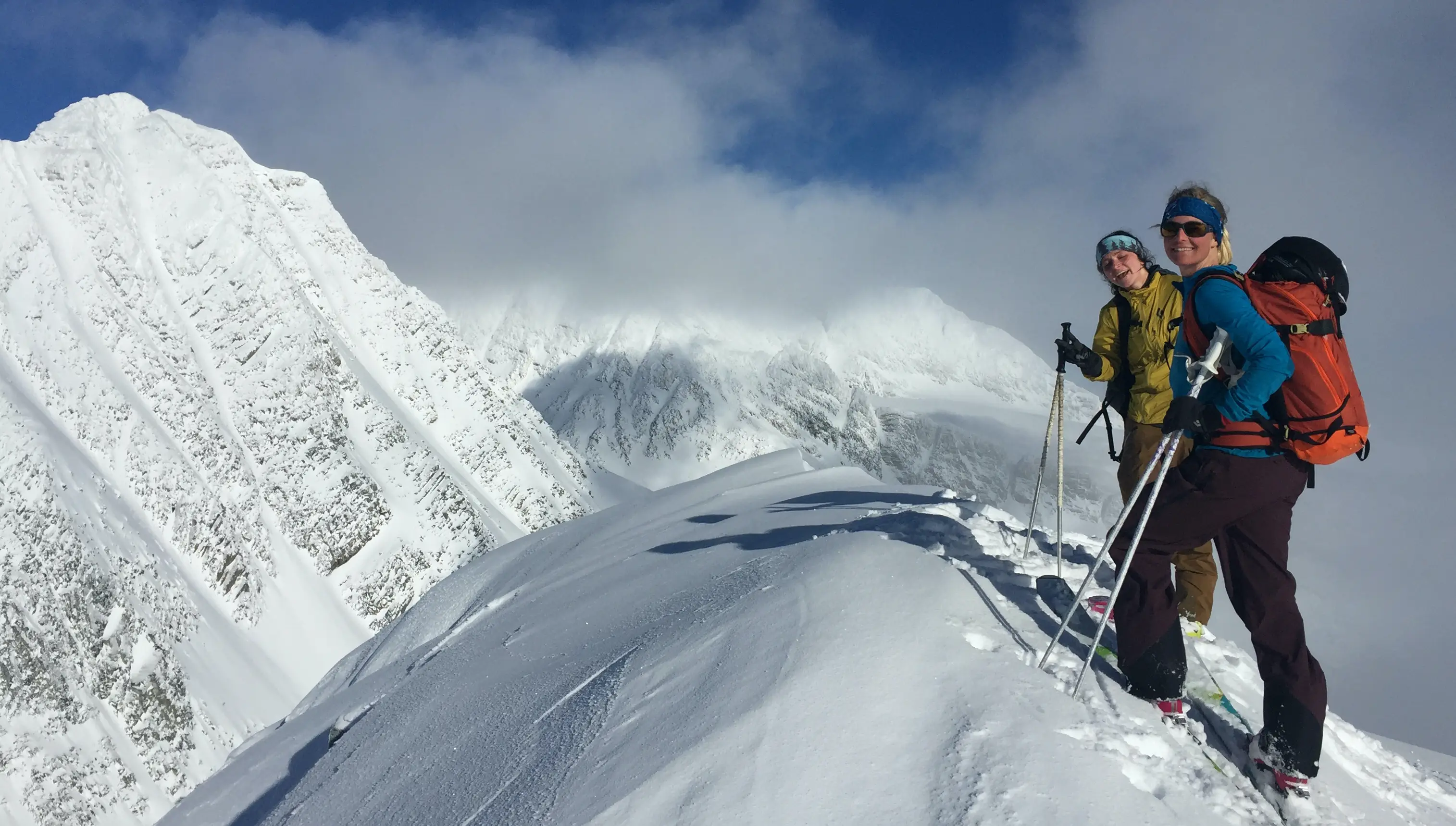 Two women stand on a snowy summit with skis on