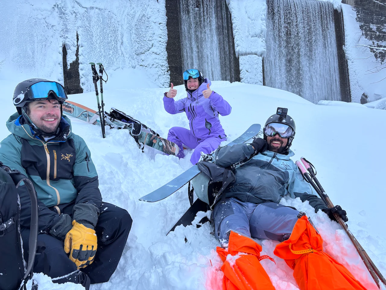 Three backcountry skiers with big smiles sitting by a waterfall in the snow