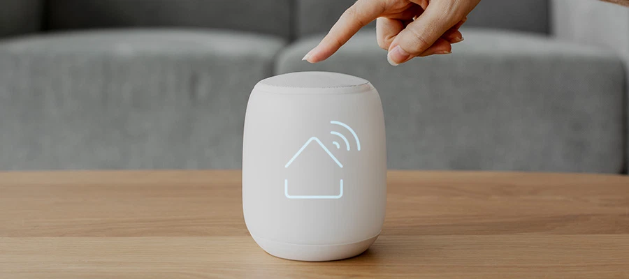 Hand tippt auf weißen Smart-Home-Lautsprecher auf Holztisch vor grauem Sofa.