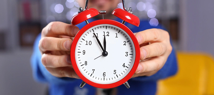Person hält einen roten Wecker, der kurz vor 12 Uhr zeigt.