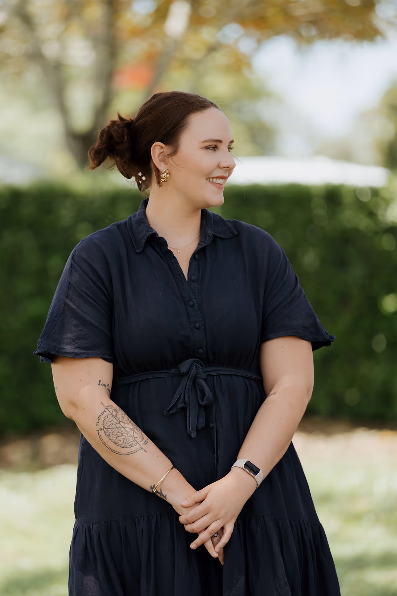 Smiling woman with dark hair tied back, wearing a black dress and showing a brain tattoo on her right forearm, standing outdoors.