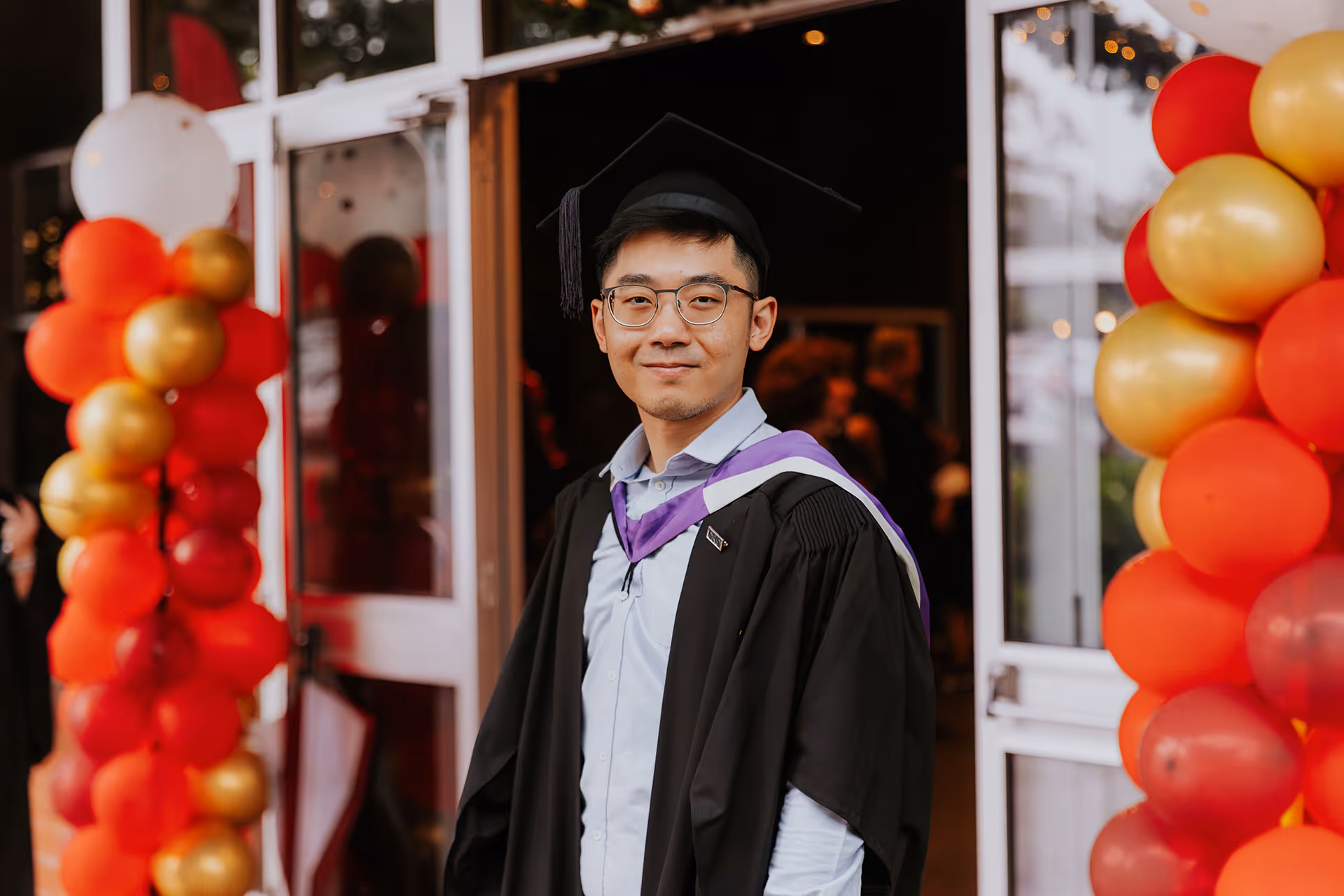 Young graduate wearing glasses and a black cap and gown standing between red and gold balloons.
