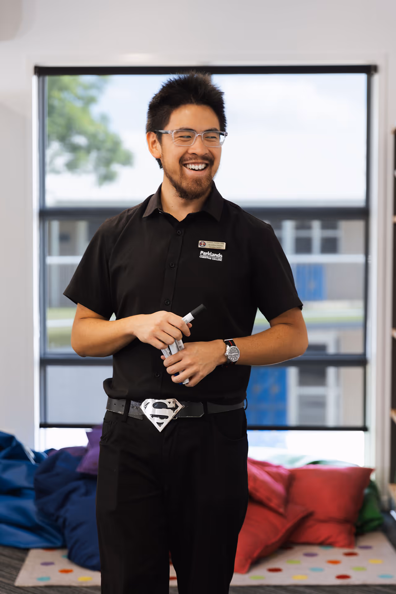 Smiling man wearing glasses and a black shirt with a name tag holding markers in a room with colorful cushions in the background.