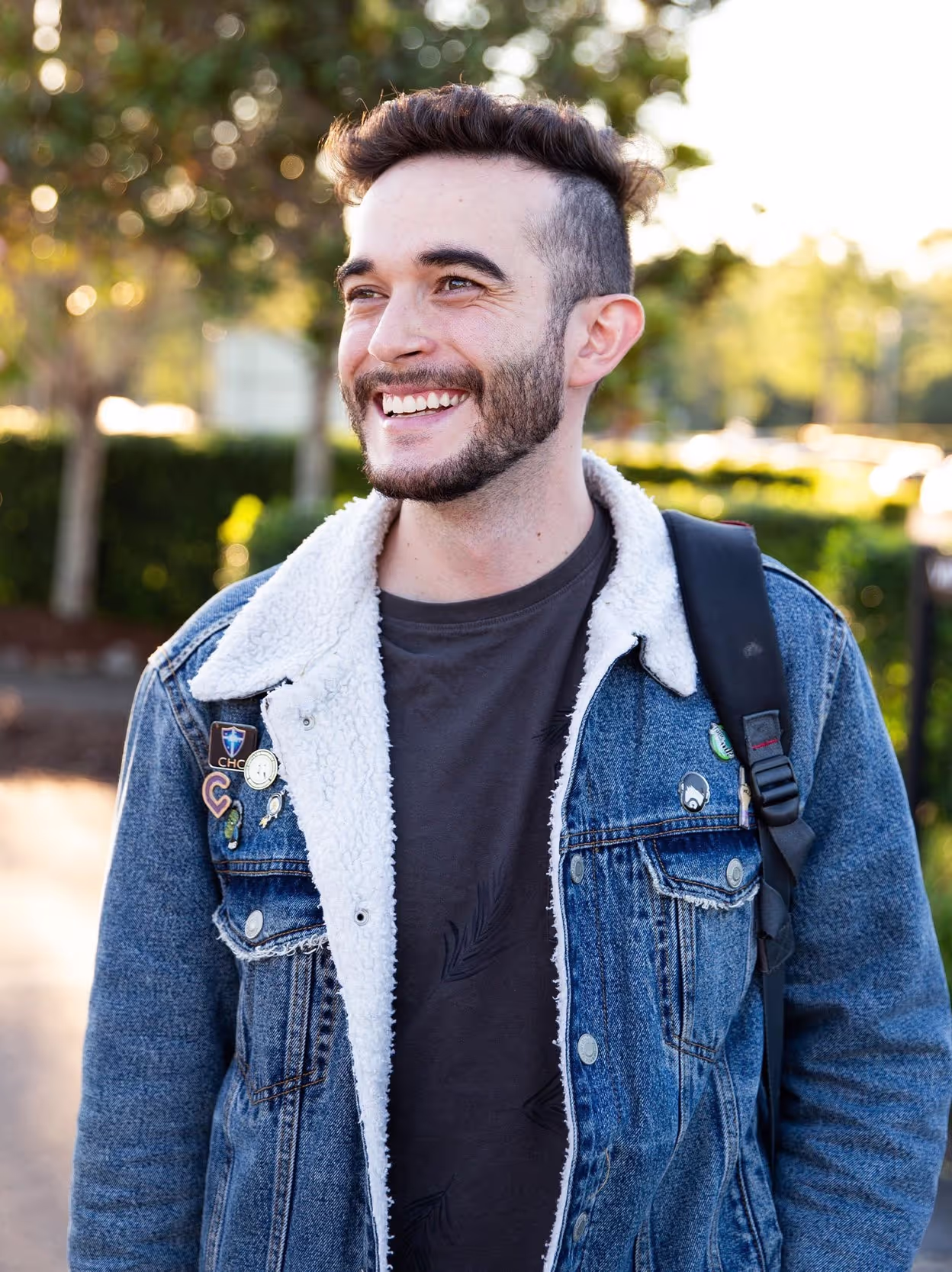 Smiling man with short dark hair and beard wearing a blue denim jacket with white fleece lining and a black backpack outdoors.