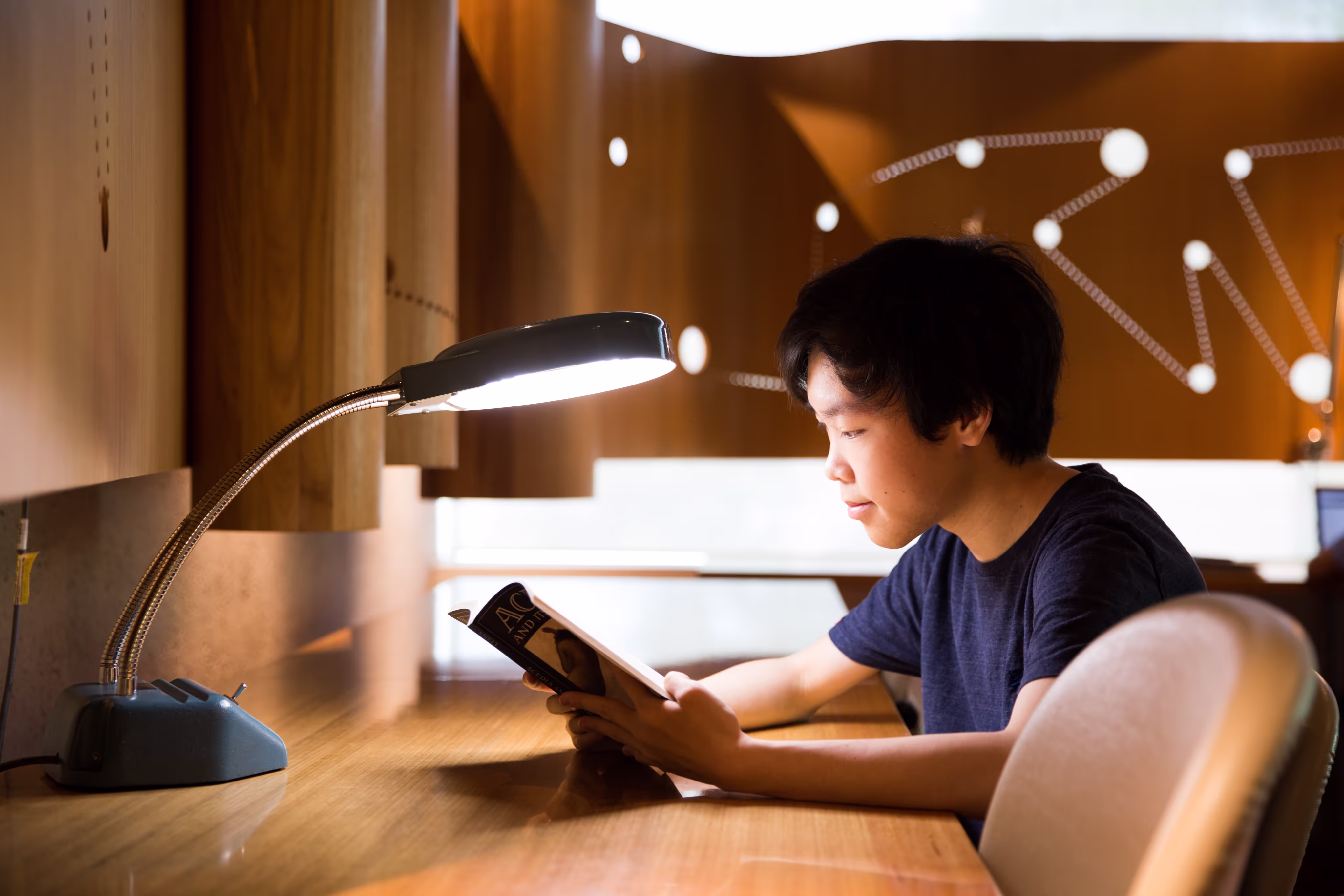 Young person with short black hair reading a book under a bright desk lamp in a wooden study area.