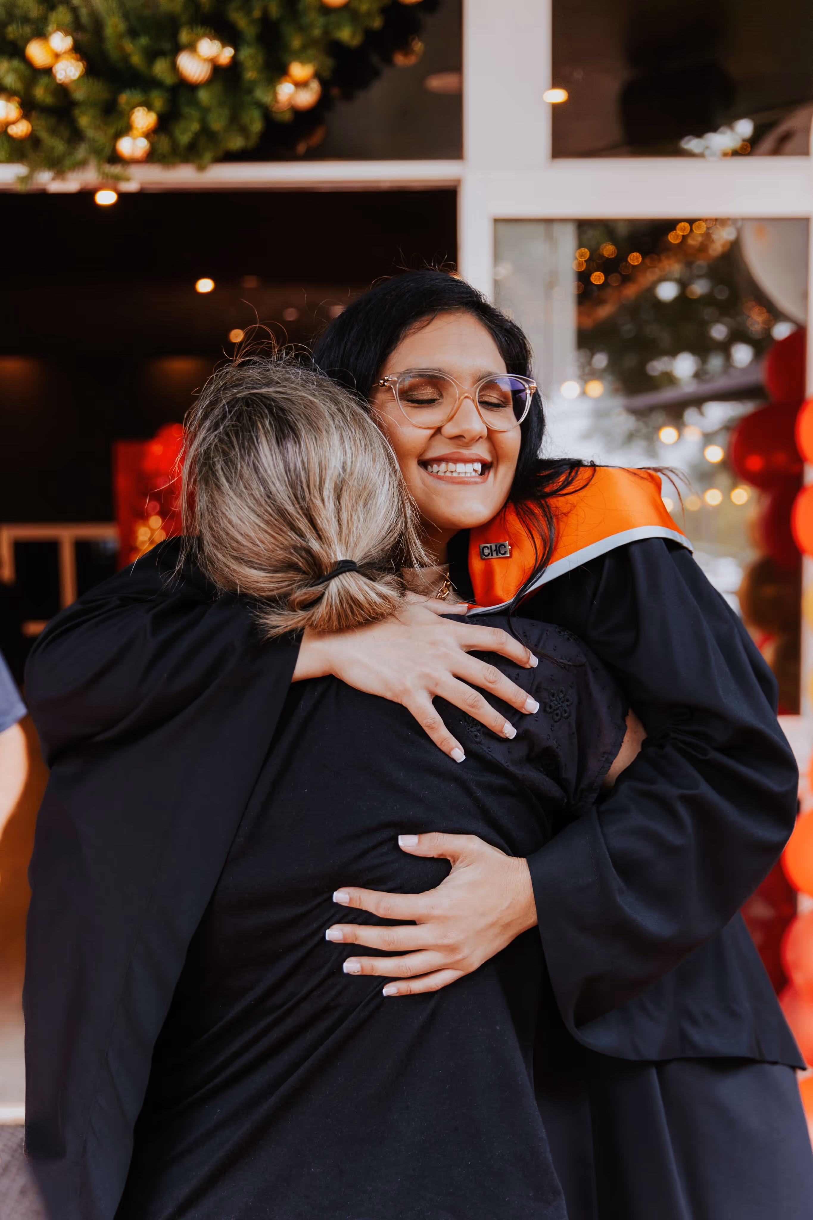 Smiling graduate in black gown and orange stole hugging a woman with blonde hair tied back.