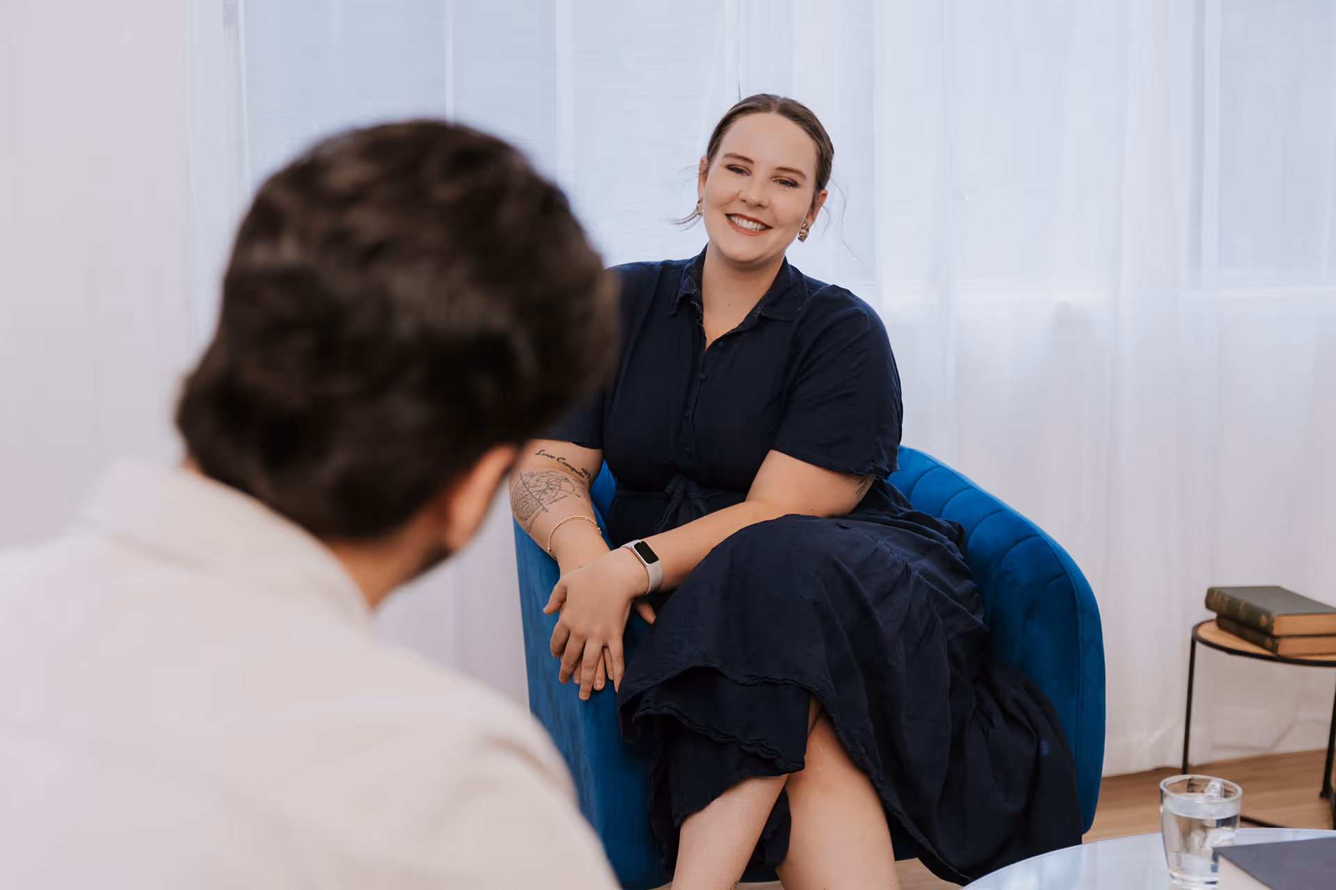 A woman in a navy dress smiling and sitting in a blue armchair while talking to a man in a white shirt.