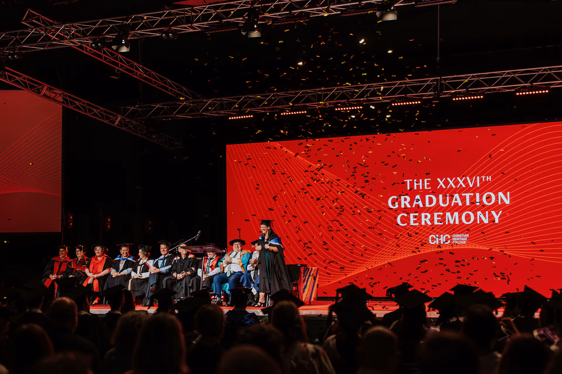 Graduates and faculty on stage at the XXXVIth Graduation Ceremony with confetti falling and a red backdrop displaying the event title.