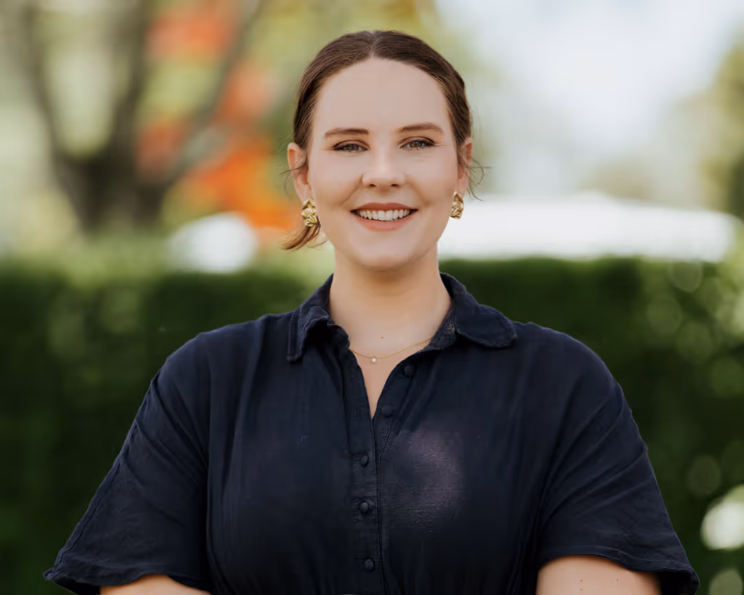 Smiling woman with brown hair, wearing a dark shirt and gold earrings, standing outdoors with blurred greenery in the background.