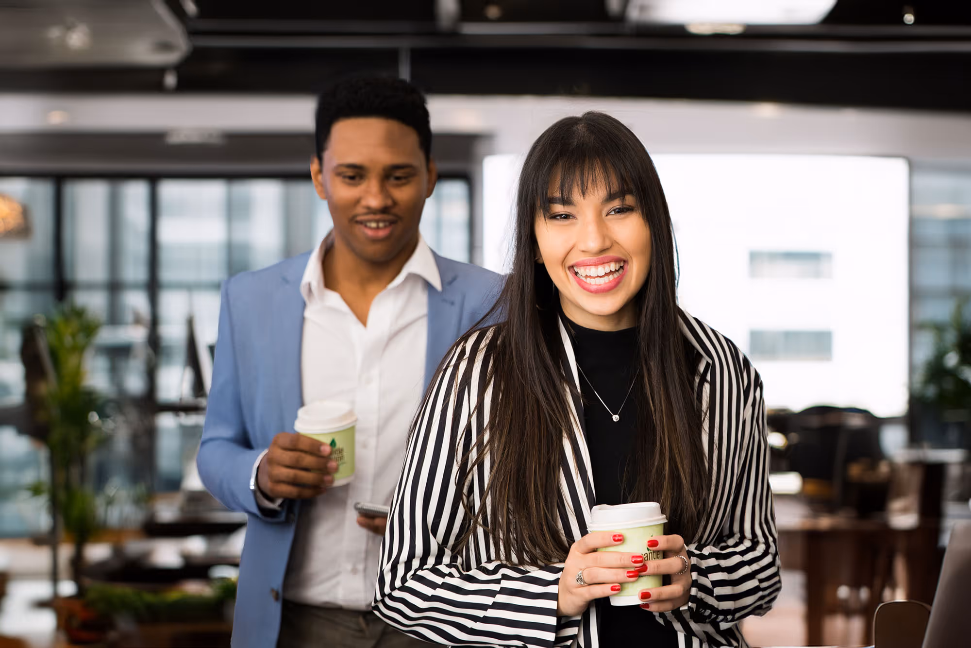 Smiling woman in a black and white striped blazer holding a coffee cup with a man in a blue blazer holding a coffee cup in the background.