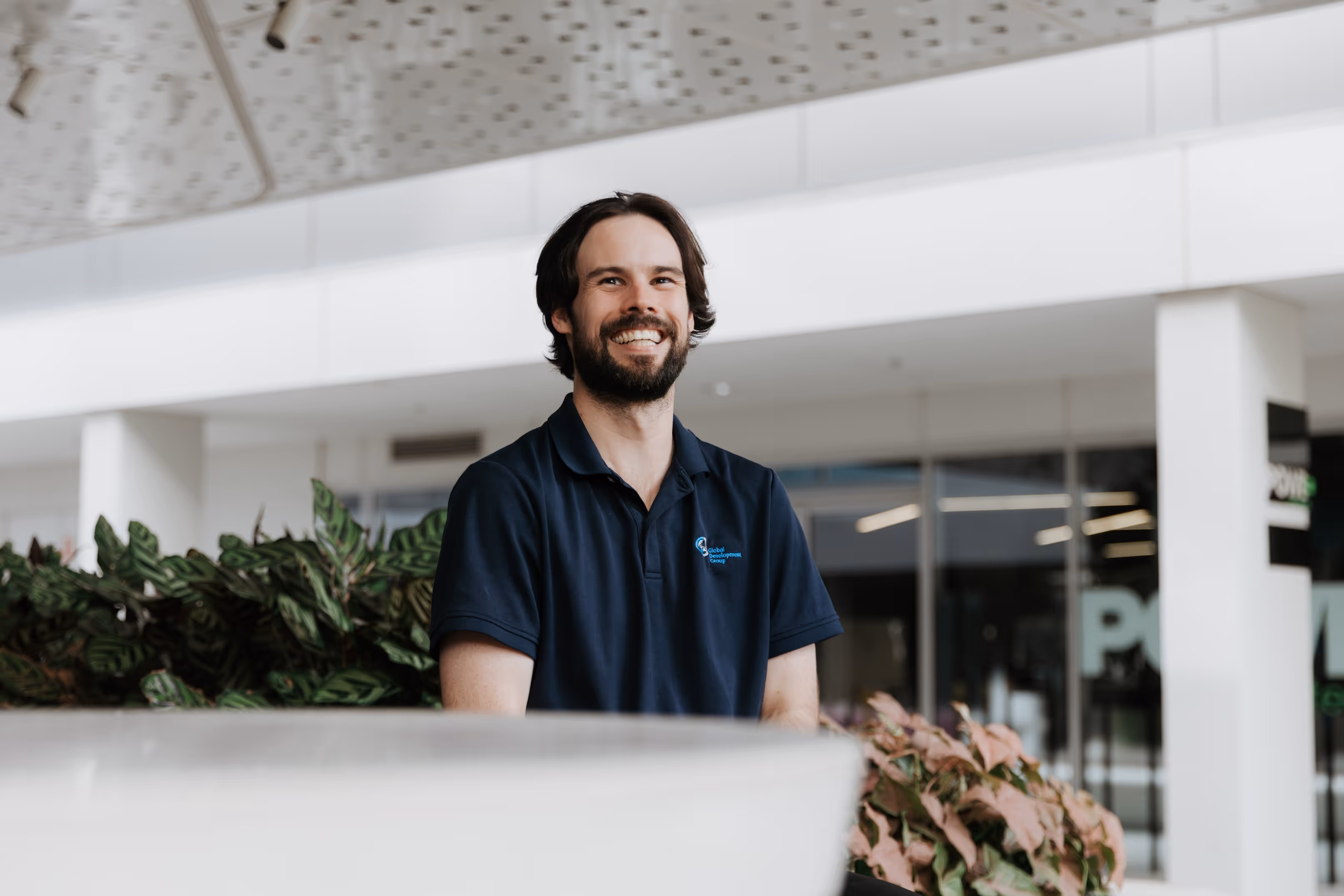 Smiling man with dark hair and beard seated indoors wearing a navy blue polo shirt with a 'Global Development Group' logo.