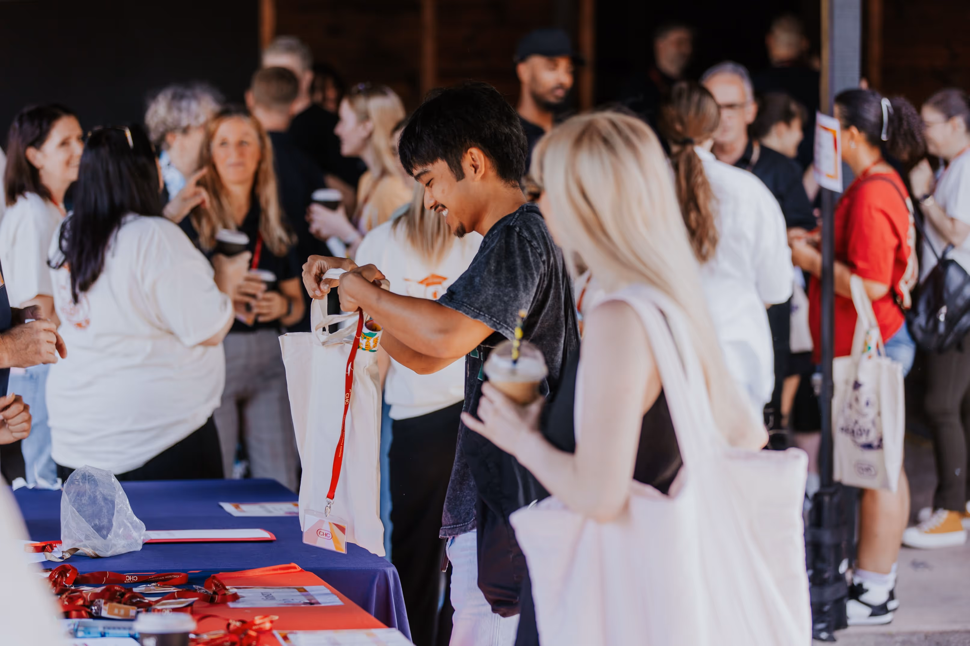People gathered at an event, with a man holding a tote bag and a woman holding a drink in the foreground.
