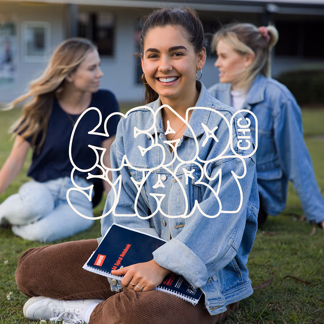 Smiling young woman in denim jacket sitting cross-legged on grass holding a notebook with two other women in denim jackets blurred in background.