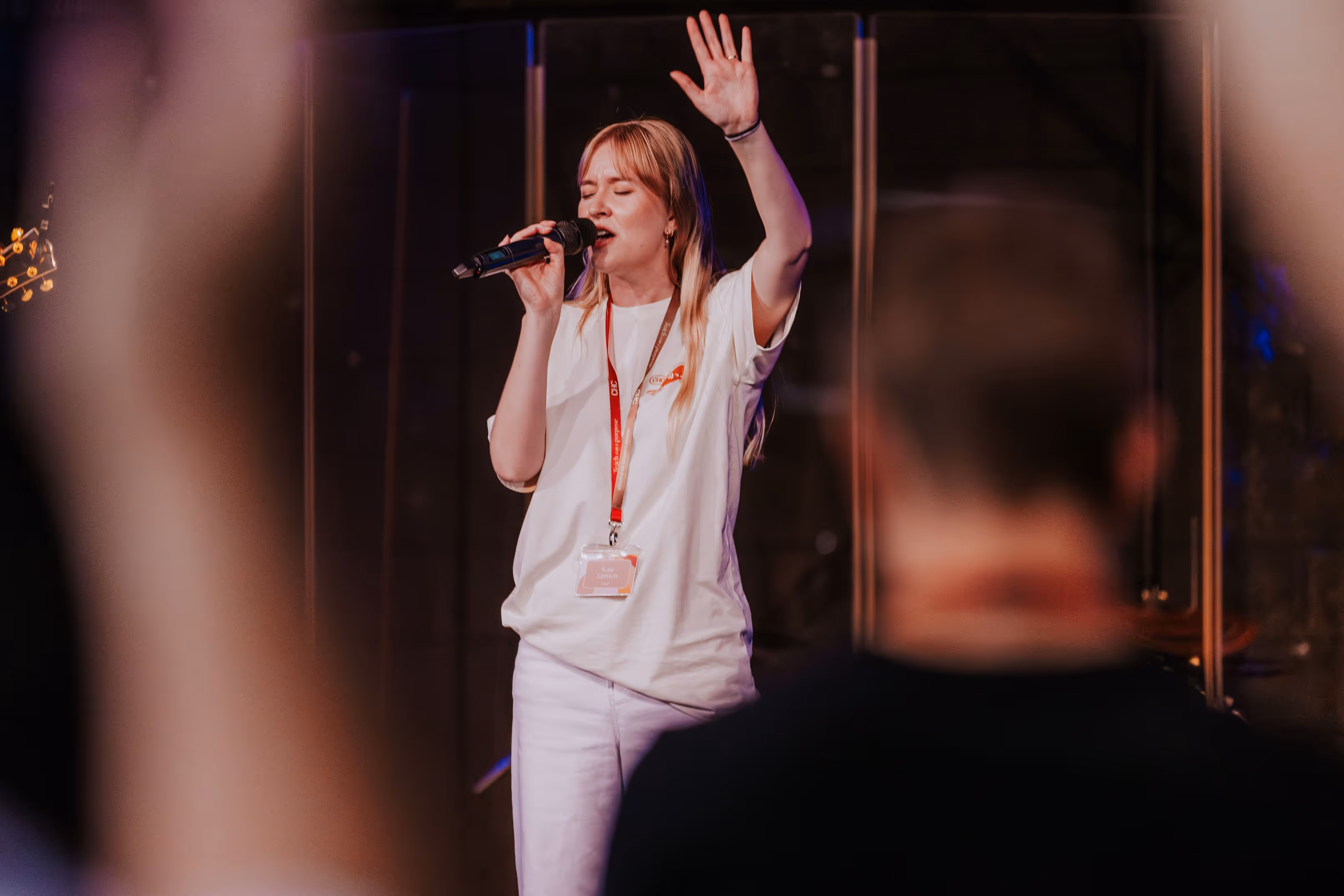 Woman with blonde hair singing into a microphone with one hand raised, wearing a white shirt and a conference lanyard.