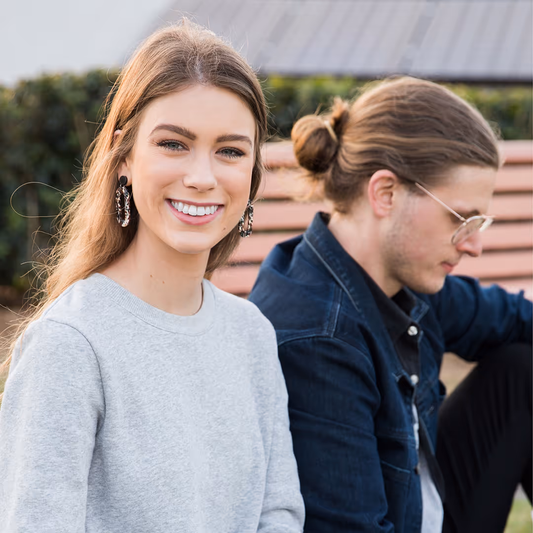 Smiling woman in a light gray sweater sitting next to a man with a bun, glasses, and a dark jacket on a bench outdoors.