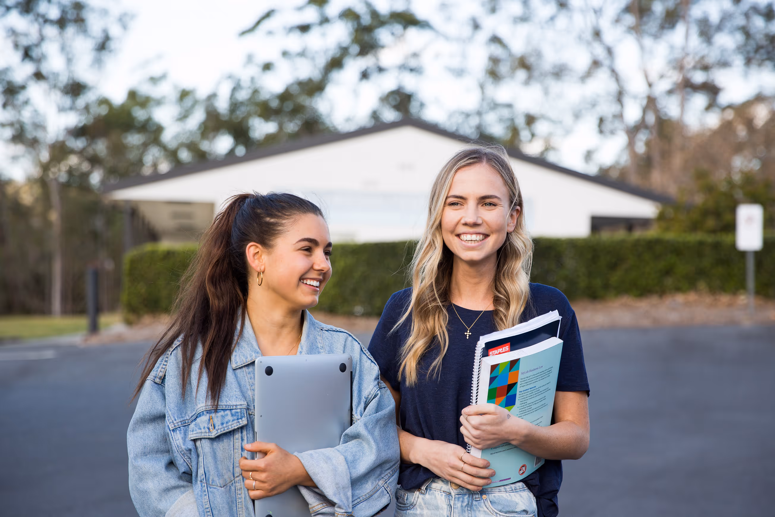 Two young women smiling and walking outdoors, one holding a laptop and the other holding notebooks and folders.