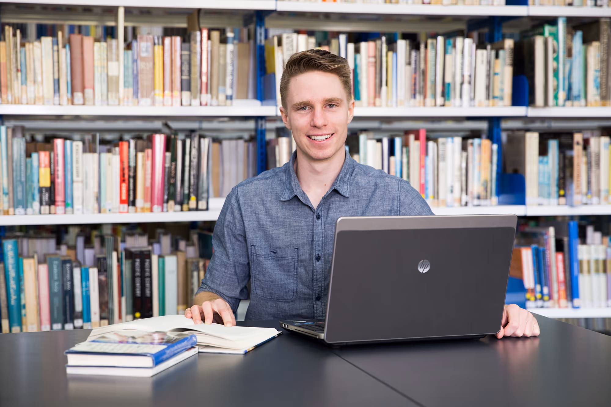 Young man smiling while sitting at a table with an open book and a laptop in a library.