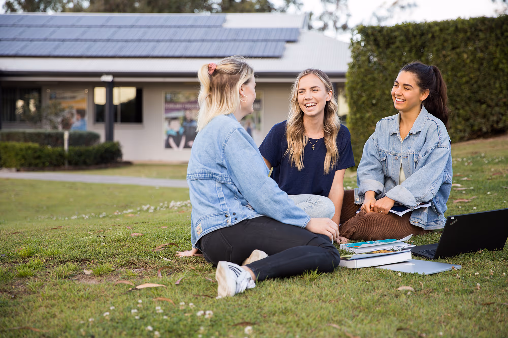 Three young women sitting on grass outside, smiling and talking with books and laptop nearby.