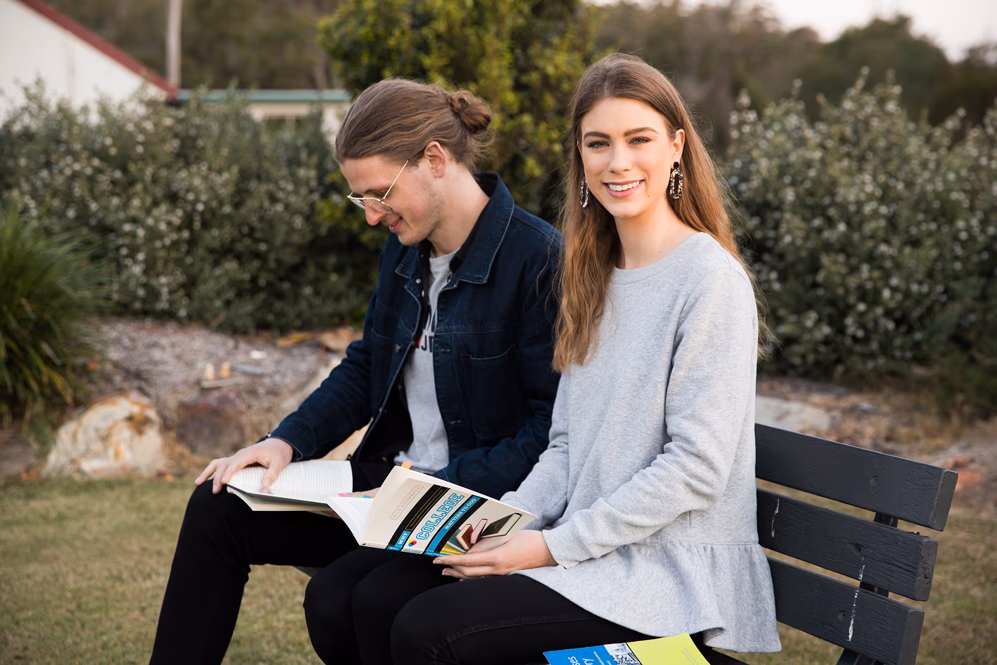 Two young adults sitting on a bench outside reading books, with one smiling at the camera.